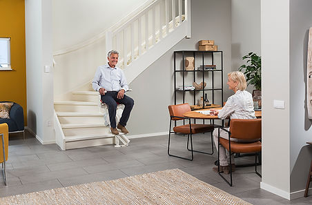Elderly man sits on stairlift; woman dines in modern home.